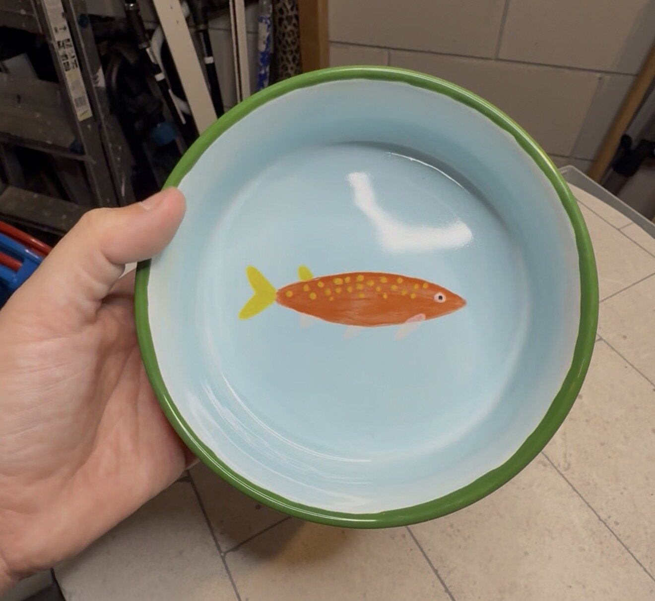 Hand met een zelf beschilderd lichtblauw keramieken bord met een oranje vis